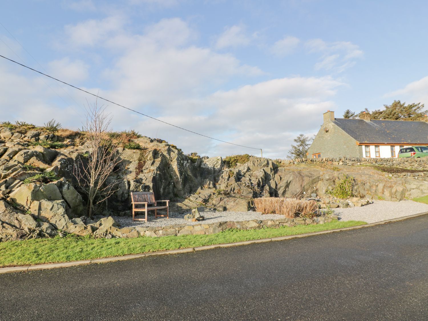 A coastal road with a rocky cliff, a bench, and a stone wall.