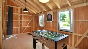 A room with wooden walls, a foosball table, a dartboard, and a window with a view of greenery.