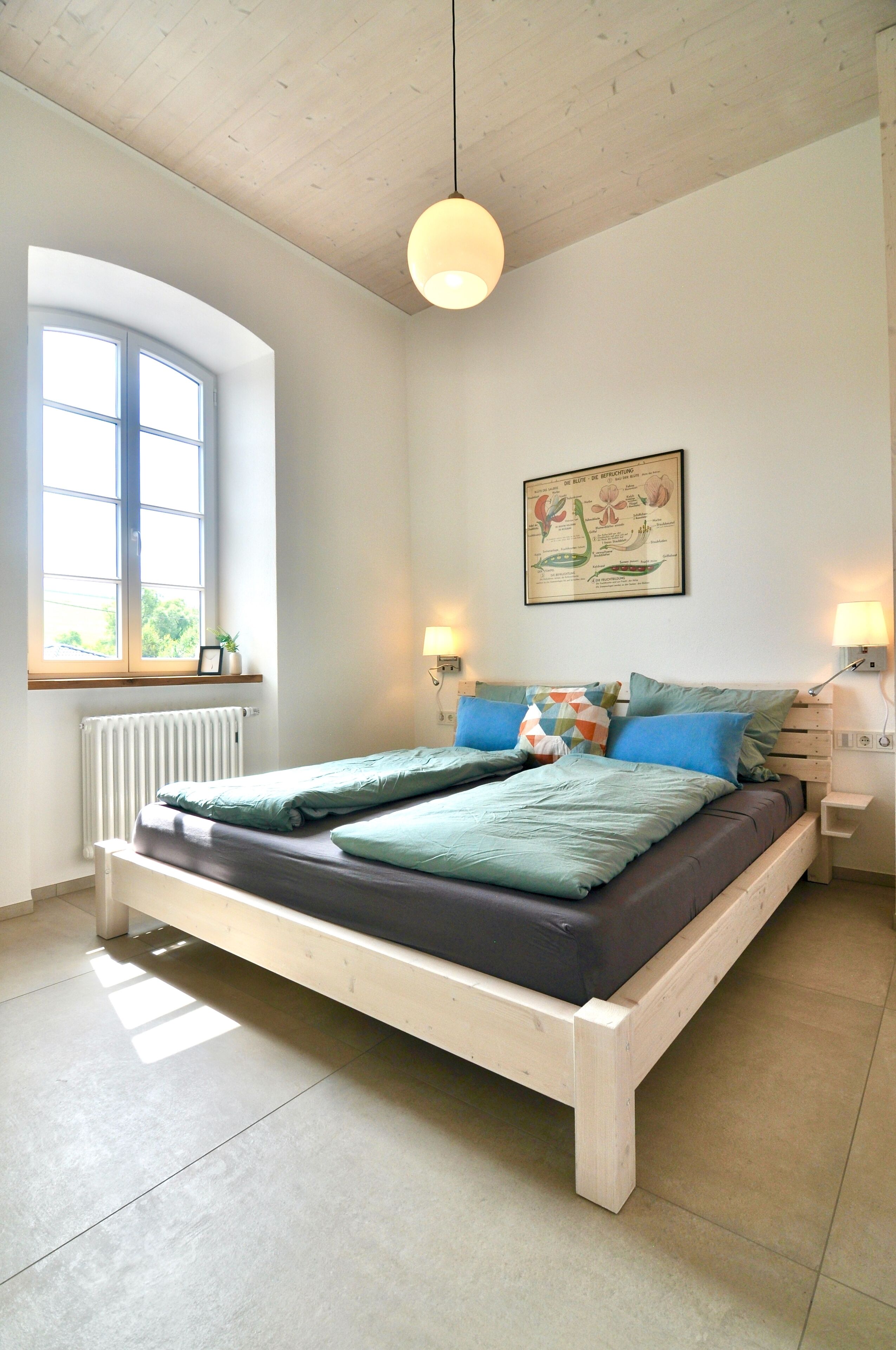 A bedroom with a wooden bed, a window with a view, a radiator, and a wall-mounted lamp.