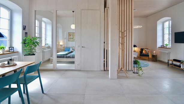 A spacious living area with a large window, a dining table with chairs, a sofa, and a wooden partition.