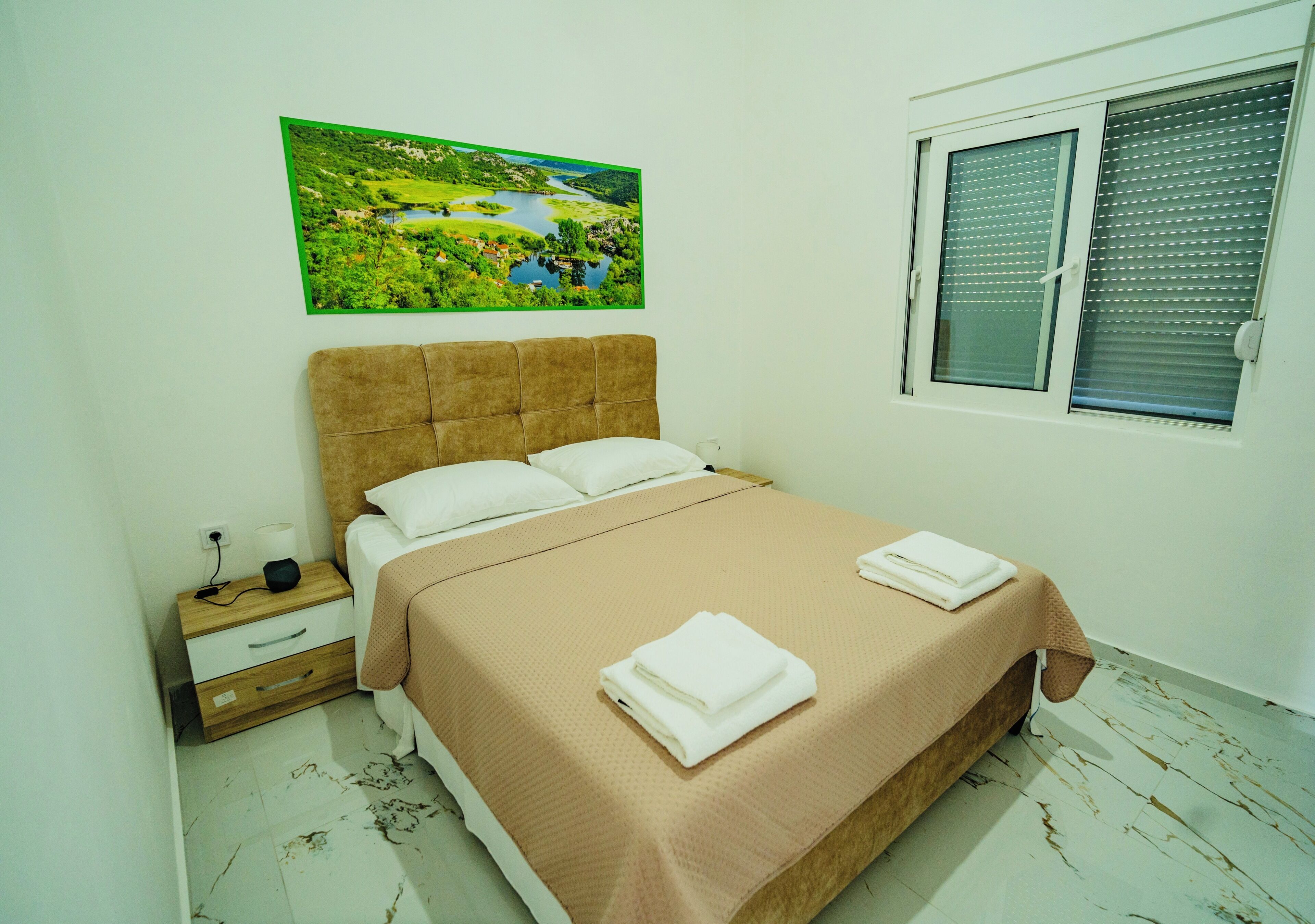 A bedroom with a bed, a nightstand, a window with blinds, and a landscape painting on the wall.
