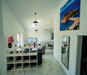 Un salon moderne avec un canapé blanc, un téléviseur et un grand tableau représentant un paysage côtier.