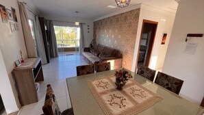 A dining area with a table set for four, a sofa in the background, and a balcony with curtains. - Your Refuge in Bombinhas – Airy family home close to the beach (Bombinhas)