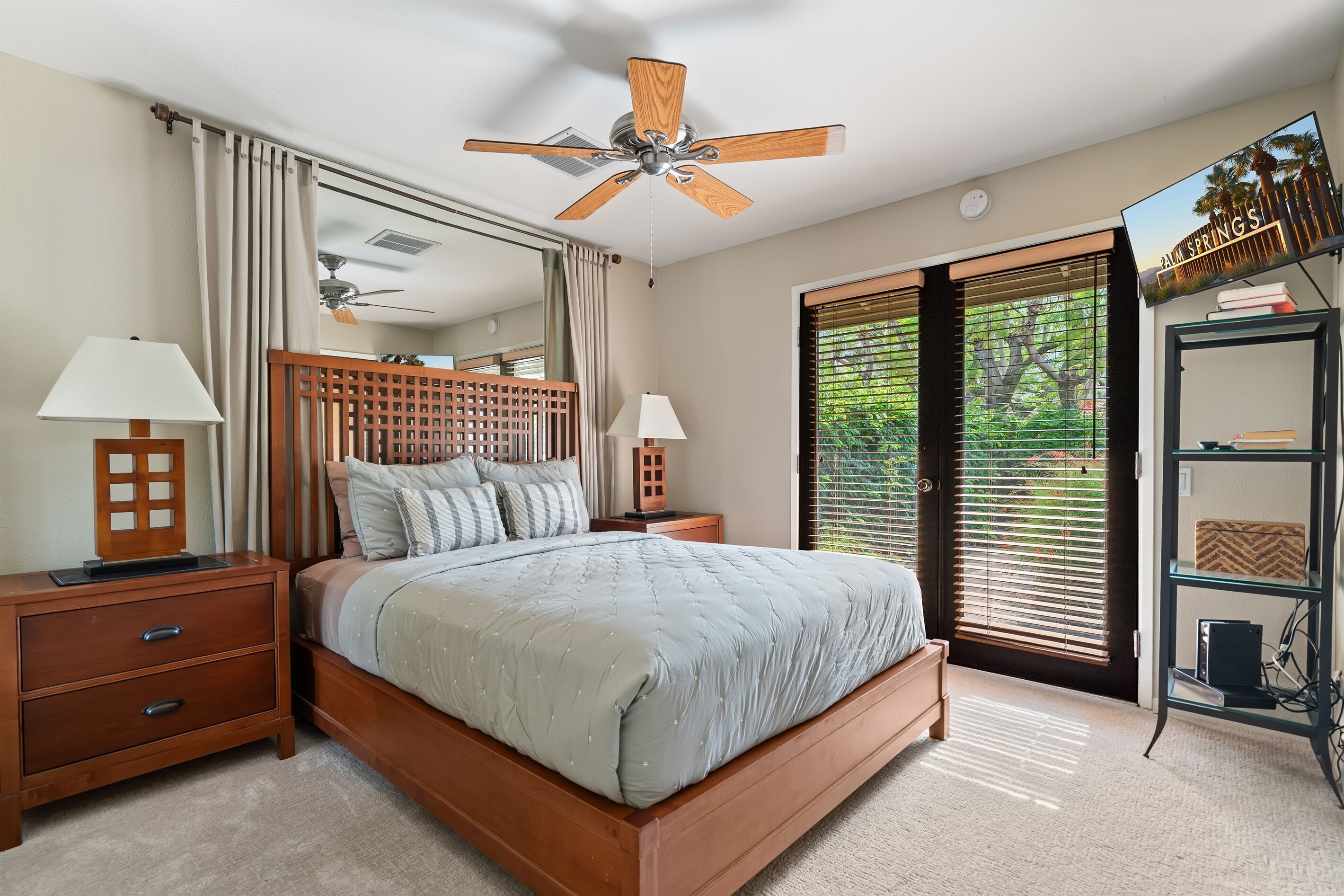 A bedroom with a bed, nightstands, a ceiling fan, a large window with blinds, and a wall-mounted TV.