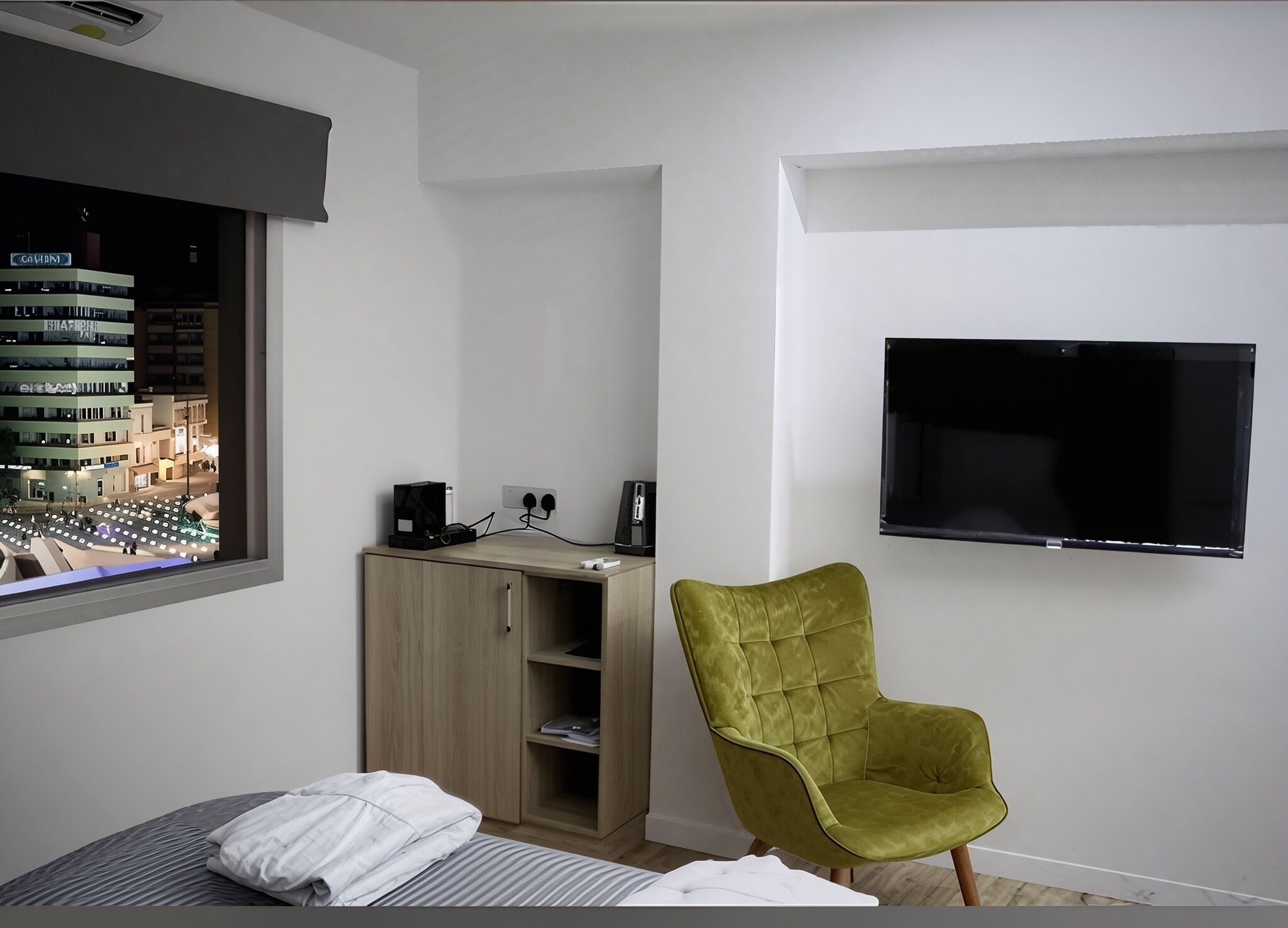 Ett sovrum med en säng, ett skrivbord med en dator, en grön stol, en tv och ett fönster med utsikt över staden.