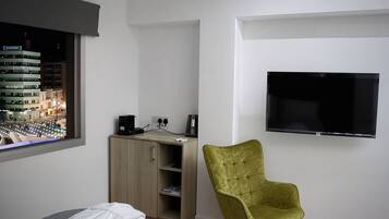 Ett sovrum med en säng, ett skrivbord med en dator, en grön stol, en tv och ett fönster med utsikt över staden.