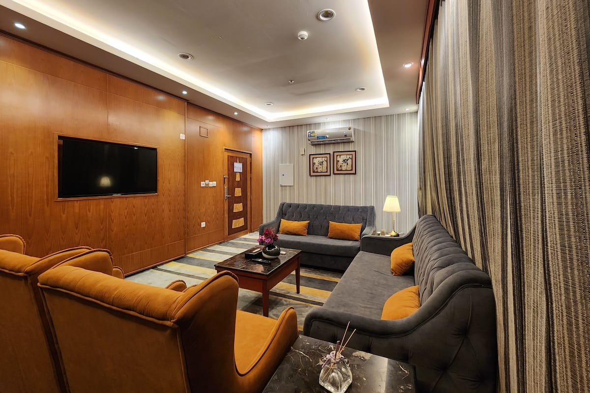 A modern hotel room with a wooden panel wall, a flat-screen TV, a sofa, two armchairs, a coffee table, and a lamp.