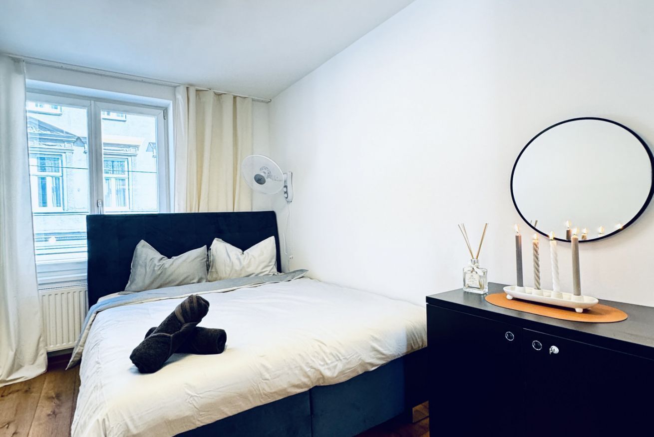 A bedroom with a bed, a bedside table with a mirror, and a window with curtains.