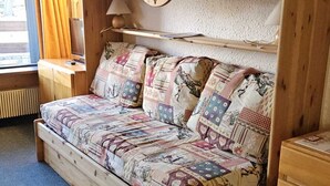 Un divano letto in legno con copripiumino patchwork, un comodino con una lampada e una finestra con tende.