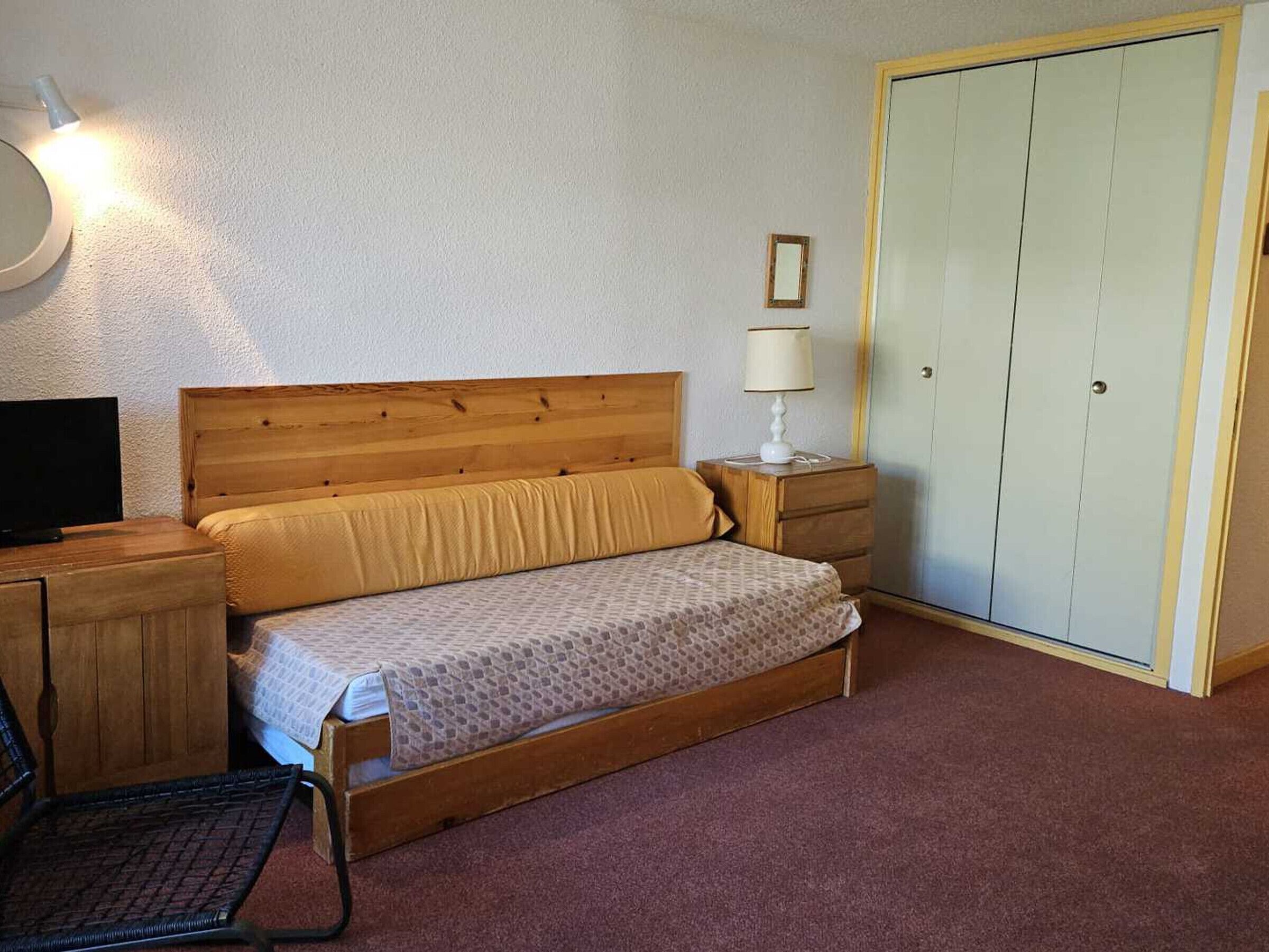 Una cama individual con cabecera de madera, una mesita de noche con lámpara, un armario y un televisor sobre un mueble de madera.