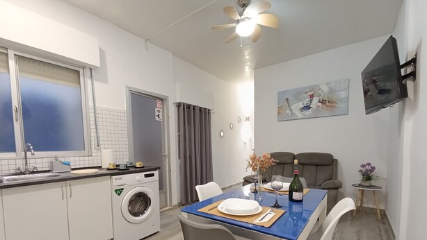 Une cuisine compacte équipée d’un lave-linge, d’une table à manger avec des chaises, d’un téléviseur fixé au mur et d’un ventilateur de plafond.