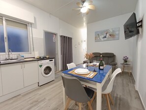 Een compacte keuken met een wasmachine, een eettafel met stoelen, een aan de muur bevestigde televisie en een plafondventilator.