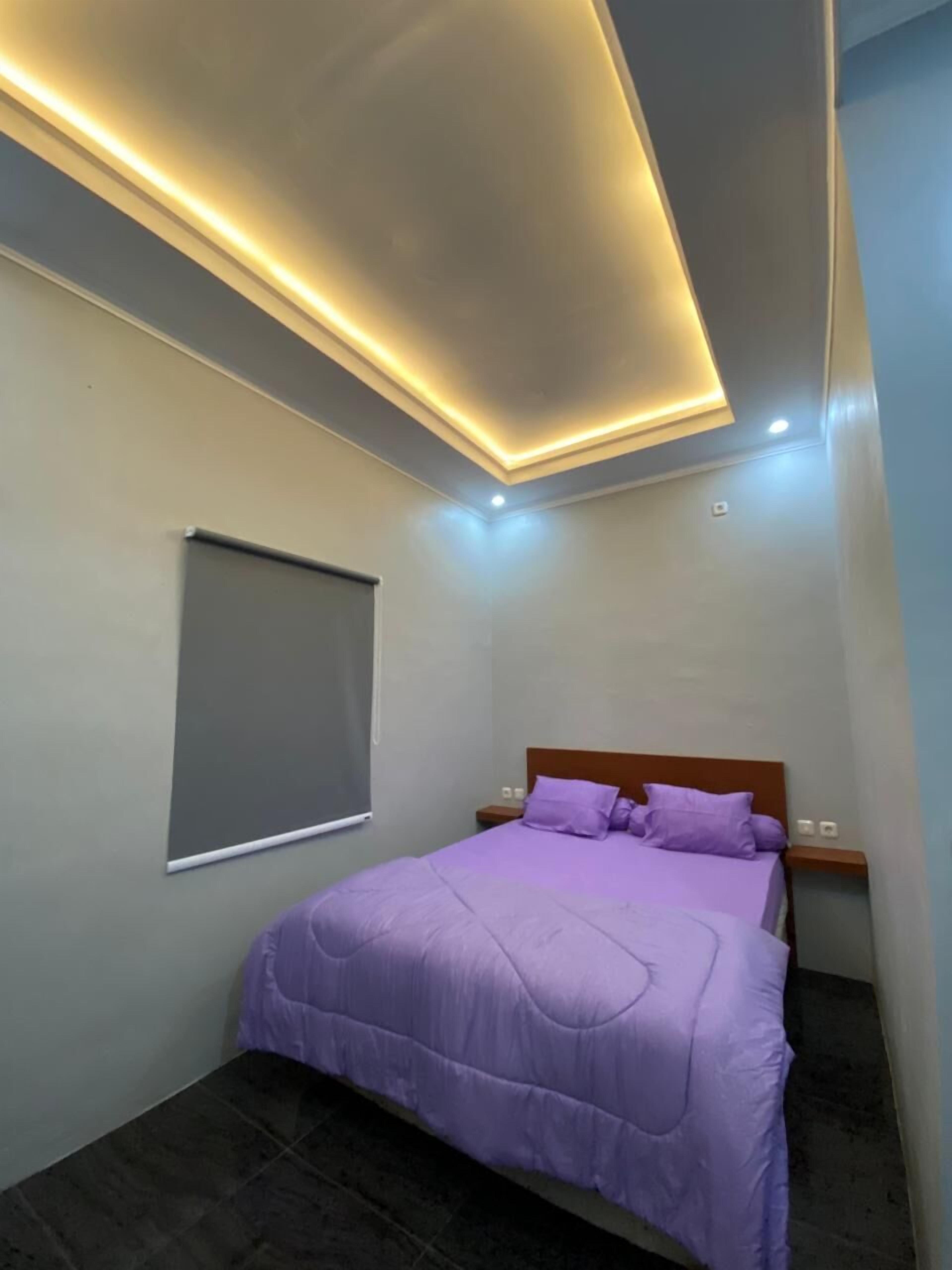 A bedroom with a bed, a nightstand, a wall-mounted shelf, and a window with a blind.