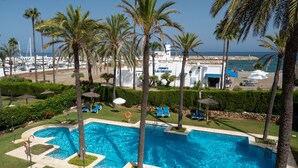 A swimming pool with palm trees, a beach, and a building with a sign. - Marina Real Port View 2457 (Manilva)