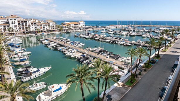 A marina with numerous boats docked, surrounded by buildings and palm trees. - Marina Real Port View 2457 (Manilva)