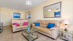 A living room with two beige sofas, one with pink and the other with blue cushions, a glass coffee table, and a large mirror on the wall. - Marina Real Port View 2457 (Manilva)