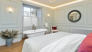 Une chambre à coucher avec un grand lit, une baignoire blanche, une fenêtre avec des rideaux, un radiateur et un miroir au mur.