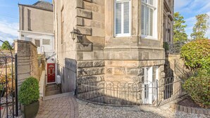 Un bâtiment en pierre avec une porte rouge, un petit jardin et une clôture en métal.
