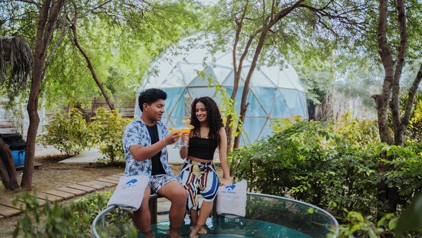 Two people sitting on a circular pool with a transparent glass edge, surrounded by lush greenery and a geodesic dome structure in the background. - Samay Playas (Playas)