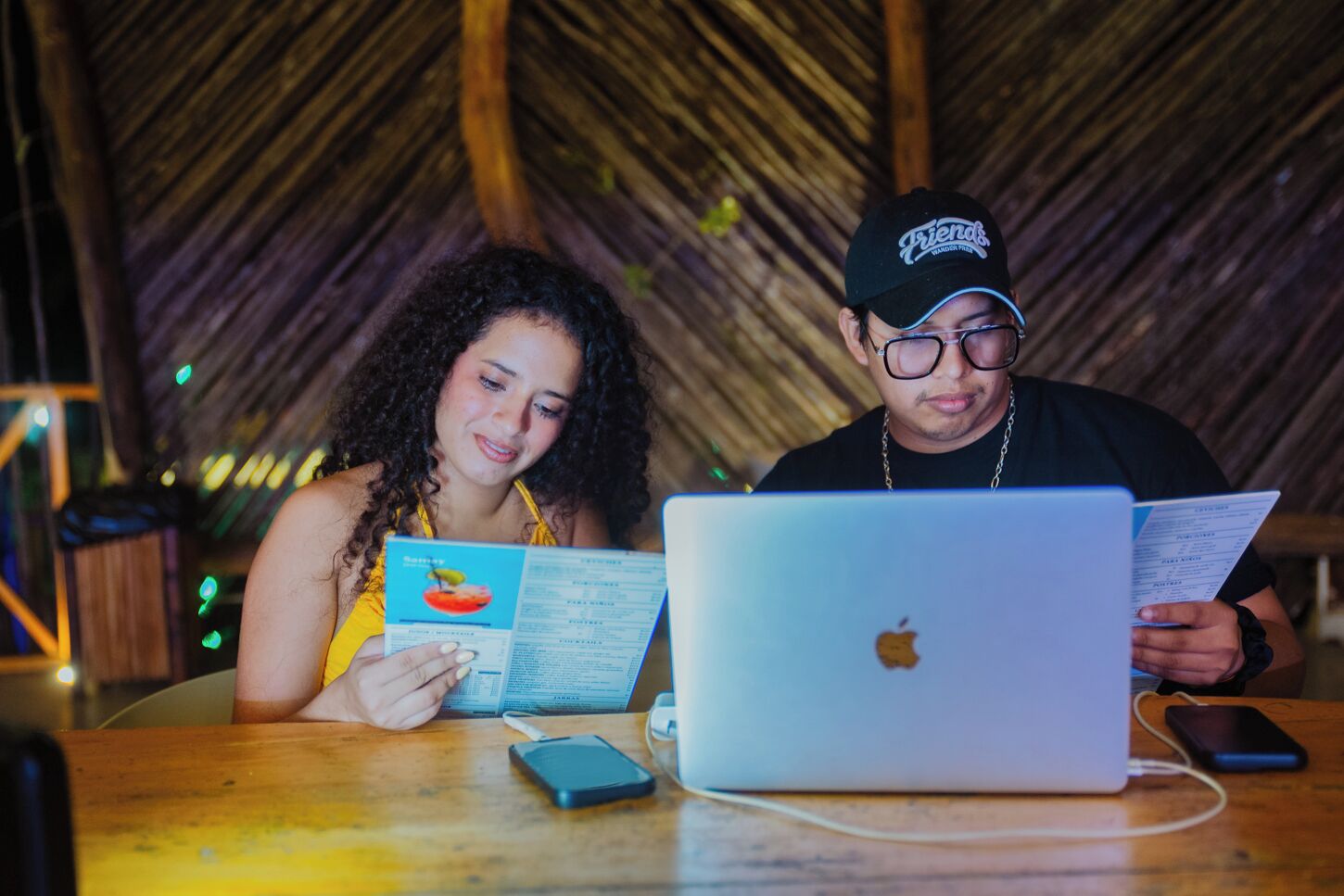 Two individuals at a wooden table with a laptop and a menu.