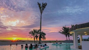 A pool area with a sunset view, palm trees, and a beach in the background.