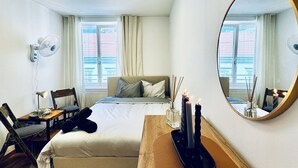 Une chambre d’hôtel comprenant un lit, une table de chevet, une chaise, une lampe, un ventilateur, un miroir et une fenêtre.