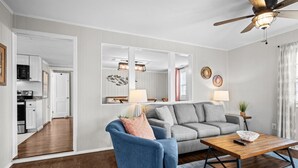 A modern living room with a blue armchair, grey sofa, and a wooden coffee table. A ceiling fan and a kitchen are visible in the background. - Bright, Beachy, and So Close to the Sea (Atlantic Beach)