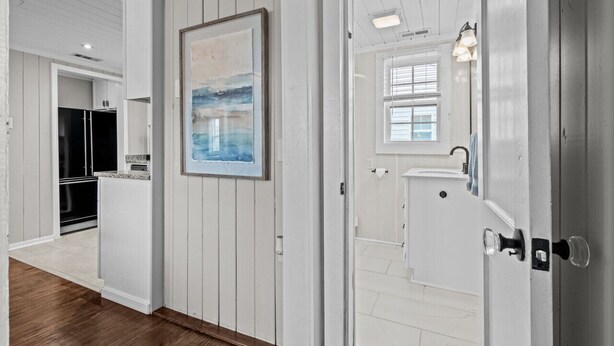 A hallway with white paneled walls, a wooden floor, and a door leading to a bathroom with a window. - Bright, Beachy, and So Close to the Sea (Atlantic Beach)