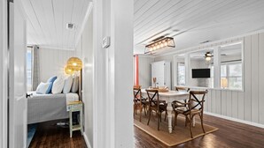 A room with a bed, bedside table, and a lamp. There is a dining area with a table, chairs, and a TV mounted on the wall. - Bright, Beachy, and So Close to the Sea (Atlantic Beach)