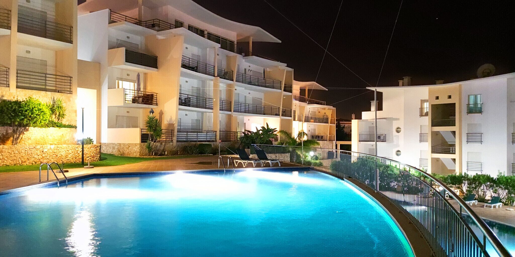 A nighttime view of a modern hotel with a swimming pool, multiple balconies, and well-lit outdoor areas.