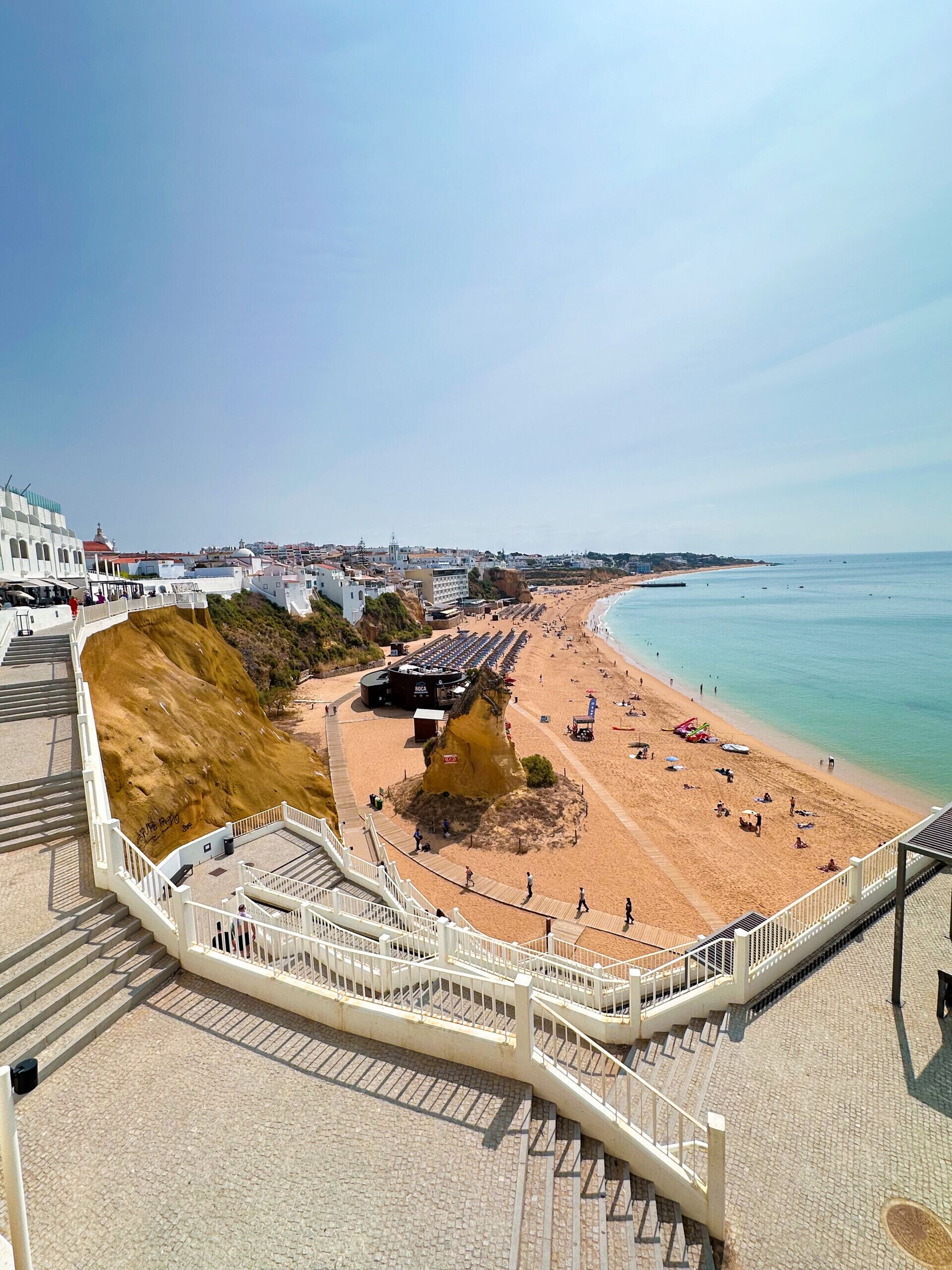 Eine Küstenpromenade mit Sandstrand, einer steilen Klippe und freiem Blick auf das Meer.