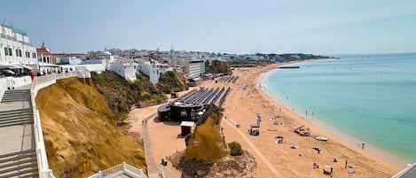 A coastal promenade with a sandy beach, a rocky cliff, and a clear view of the sea.