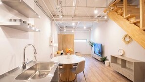 Une cuisine moderne avec un évier, une table à manger et des chaises. On aperçoit un escalier et un téléviseur au fond.