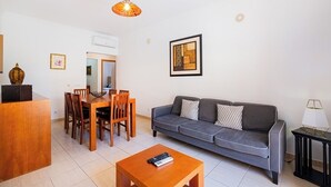 A modern living room with a grey sofa, wooden dining table, and chairs. There is a small table with a lamp and a framed picture on the wall.