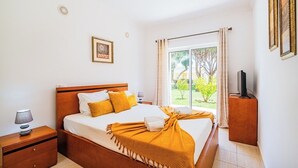 A bedroom with a large bed, a nightstand, a TV stand, and a window with curtains.