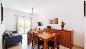 A dining area with a wooden table and chairs, a sofa, a TV on the wall, and a cabinet with decorative items.