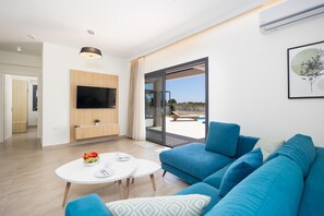 A modern living room with a blue sofa, a flat-screen TV mounted on the wall, a round white coffee table, and a view of a pool through a glass door.