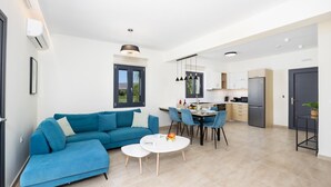 A modern living room with a blue sofa, a dining table with chairs, and a kitchen area with a refrigerator and cabinets. - Villa Pinelopi Tria - Two Bedroom Villa, Sleeps 5 (Ionian Islands)