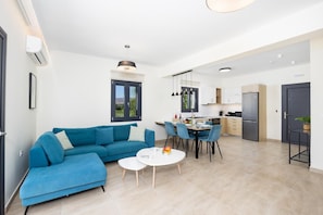 A modern living room with a blue sofa, a dining table with chairs, and a kitchen area with a refrigerator and cabinets.