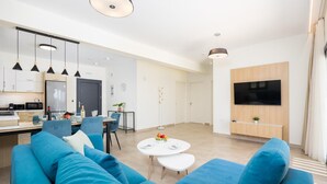 A modern living room with a blue sofa, a dining area with a table and chairs, a kitchen with a microwave, and a TV mounted on the wall. - Villa Pinelopi Tria - Two Bedroom Villa, Sleeps 5 (Ionian Islands)