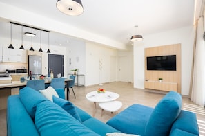 A modern living room with a blue sofa, a dining area with a table and chairs, a kitchen with a microwave, and a TV mounted on the wall.