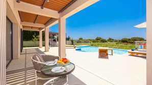 A covered outdoor area with a pool, lounge chairs, and a table with fruit and cutlery. - Villa Pinelopi Tria - Two Bedroom Villa, Sleeps 5 (Ionian Islands)
