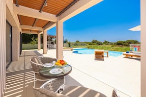 A covered outdoor area with a pool, lounge chairs, and a table with fruit and cutlery.
