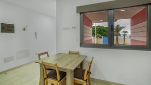 A room with a wooden table and chairs, a window with a view of greenery and buildings, and a wall-mounted light switch.