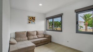 A room with a beige sectional sofa, a window with a view of palm trees, and a painting on the wall.