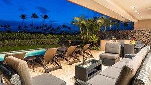 A poolside area with wicker furniture, a stone wall, and a view of palm trees against a twilight sky. - Laule‘a #13 by Gather – Mauna Lani 4BR Villa w/ Golf Frontage, Private Pool/Spa (Kamuela)
