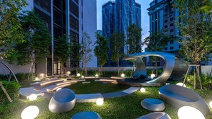 A modern outdoor seating area with circular benches and a unique curved structure, surrounded by greenery and tall buildings.
