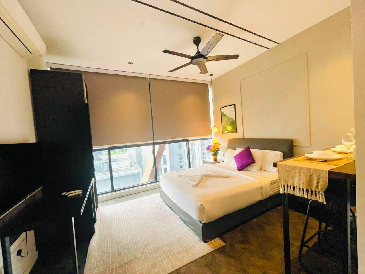 A hotel room with a bed, a desk, a chair, a ceiling fan, and a window with a view of buildings.