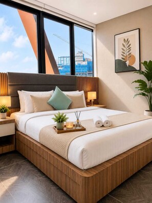 Una habitación de hotel moderna con una cama grande, cabecera de madera, dos mesitas de noche con lámparas, una ventana con vistas a una obra en construcción y una obra de arte enmarcada en la pared.