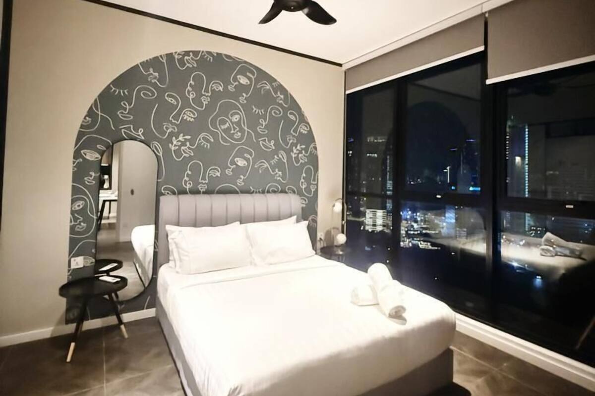 A modern bedroom with a large bed, a ceiling fan, a view of the city at night, and a unique wall design.